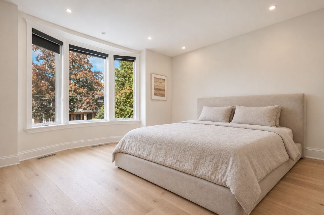 Primary bedroom with a three-panel bay window, white oak flooring and treetop views
