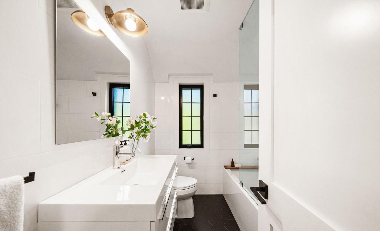Main bathroom with large-format white wall tile, black hexagon floor tile, tub with glass screen and brass lighting