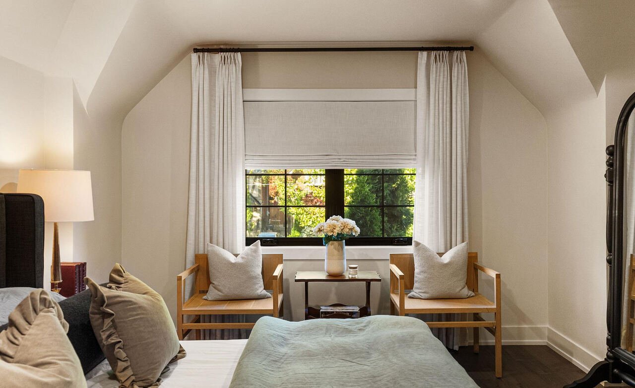 Primary bedroom under a sloped ceiling with dormer window, linen drapery and a pair of oak armchairs